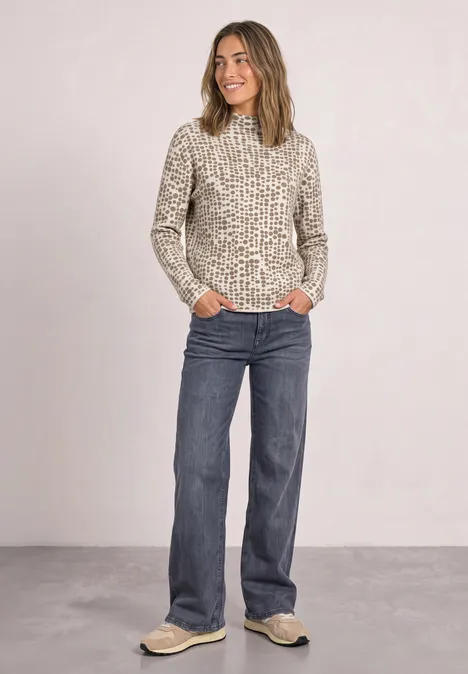 Frau in Pullover mit Tierprint, blauen Jeans und Sneakers steht entspannt vor neutralem Hintergrund.