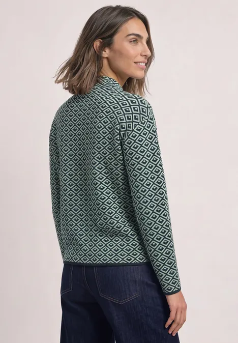 Frau steht seitlich im grünen, gemusterten Strickpullover und schaut über die Schulter.