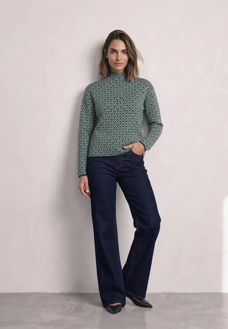 Frau in grün gemustertem Pullover und weiten dunkelblauen Jeans vor hellem Hintergrund.