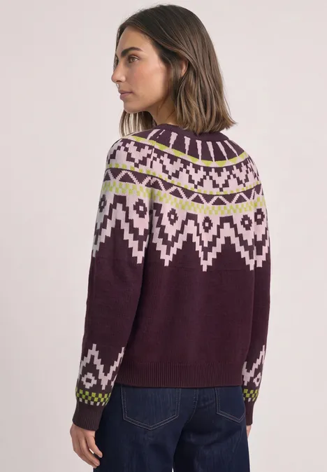 Frau trägt violetten Norwegerpullover mit weiß-grünem Muster, steht seitlich in Jeans vor hellem Hintergrund.