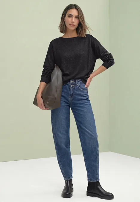 Frau in schwarzem Sweater, blauen High-Waist-Jeans, schwarzen Stiefeletten, schwarze Tasche vor hellem Hintergrund.