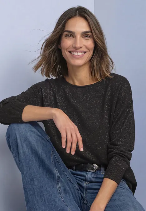 Eine lächelnde Frau mit braunen Haaren in dunkel meliertem Pullover und blauen Jeans sitzt entspannt.