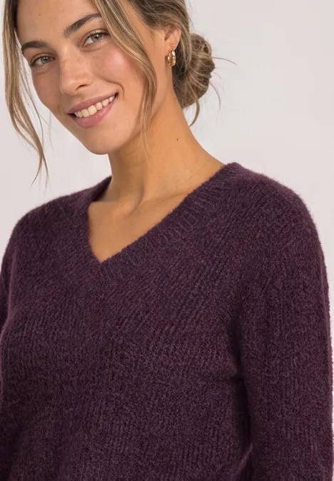 Frau mit blondem Dutt trägt violetten Strickpullover mit V-Ausschnitt und lächelt.