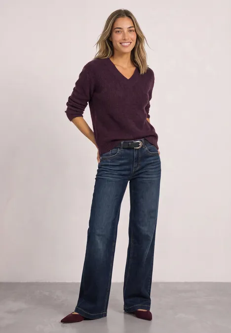 Frau in dunkler Jeans und lila Pullover vor einfarbigem Hintergrund.