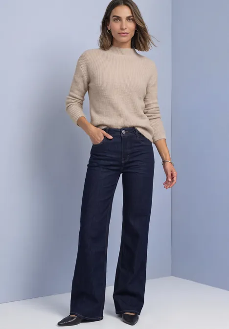 Frau in beigem Pullover und weitem, hoch tailliertem dunkelblauen Jeans vor hellem Hintergrund.