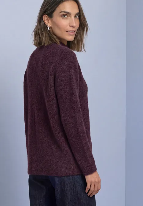Frau in dunkelrotem Strickpullover und Jeans vor neutralem Hintergrund, blickt über die Schulter.