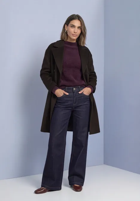 Eine Frau steht vor pastellblauem Hintergrund in Mantel, Pullover und weiten Jeans.