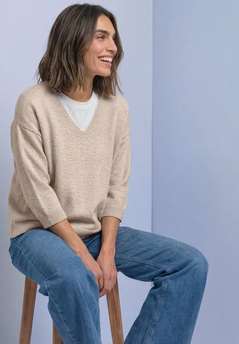 Eine lächelnde Frau auf einem Hocker trägt beigen V-Pullover, weißes Shirt und Jeans vor hellblauem Hintergrund