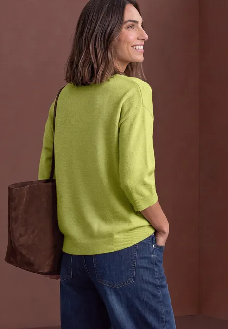 Une femme sourit de côté, vêtue d'un pull vert, d’un jean et portant un sac sur l’épaule, fond neutre.