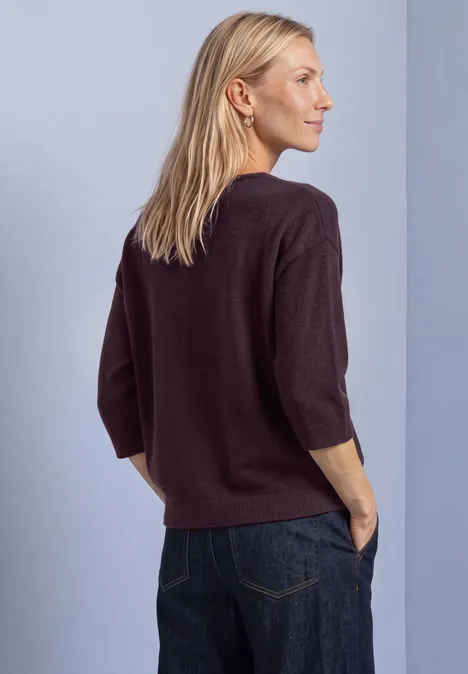Frau mit lockerem, dunkelviolettem 3/4-Ärmel-Pullover und Jeans, seitlich stehend, Hände in den Taschen.