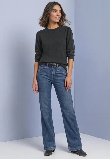 Pullover mit Rundhals und Glitzertape armor grey mel. Frau in schwarzem Pullover, blauen Jeans und schwarzen Schuhen steht vor schlichter grauer Wand und lächelt.