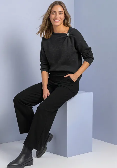 Pullover mit Boat Neck und Broschendetail armor grey mel. Eine lächelnde Frau in dunkelgrauem Pullover, schwarzer weiter Hose und Stiefeln sitzt auf einem hellen Würfel.