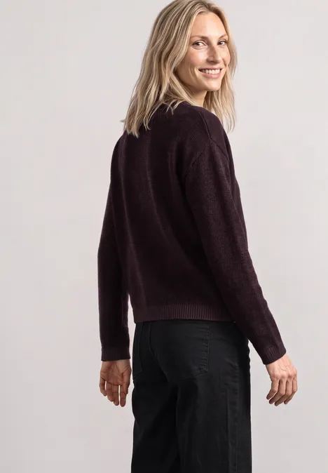Frau mit blondem Haar trägt dunkelroten Pullover und schwarze Hose, schaut über die Schulter und lächelt.