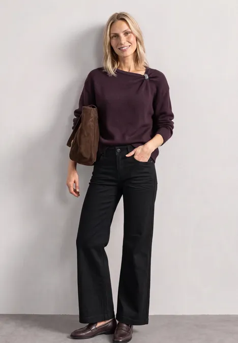 Eine lächelnde Frau mit blondem Haar trägt einen lila Pullover, schwarze weite Hosen und eine braune Tasche.