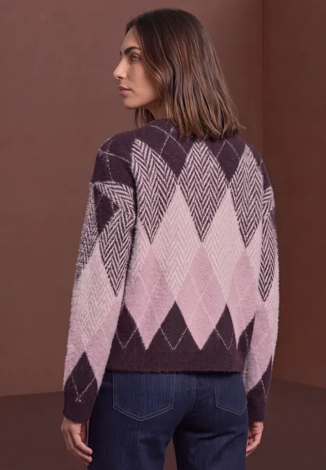 Frau steht seitlich, trägt violett-rosa Argyle-Pullover und Jeans vor braunem Hintergrund.