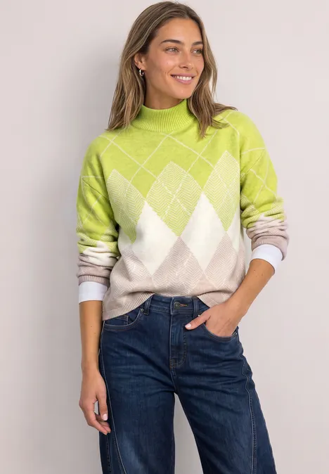 Junge Frau in grün-weißem Rautensweater und Jeans, lächelnd mit Hand in Tasche.