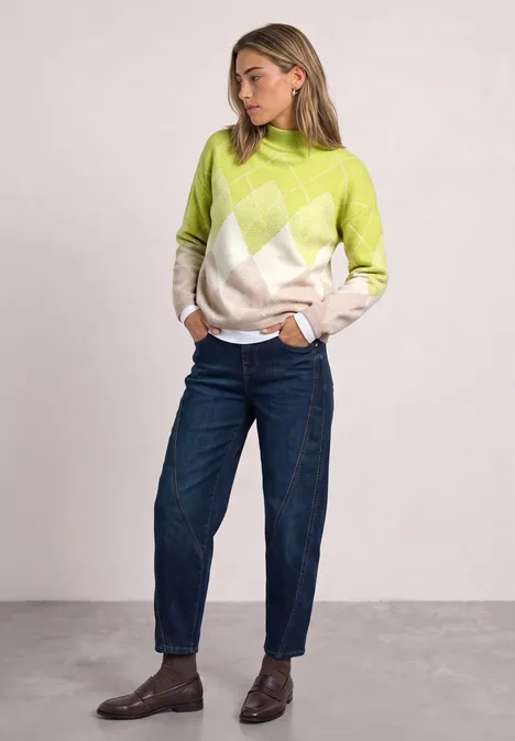 Frau in lockerem, argyle-muster Pullover, dunkle Jeans und braune Boots, steht vor neutralem Hintergrund.