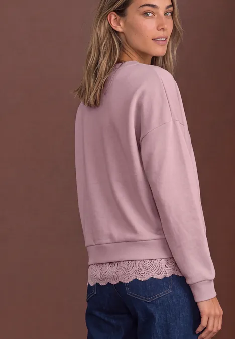 Sweat-shirt ample avec bordure en dentelle quiet rose Frau von hinten in rosa Sweatshirt mit Spitzenabschluss über blauen Jeans vor braunem Hintergrund.