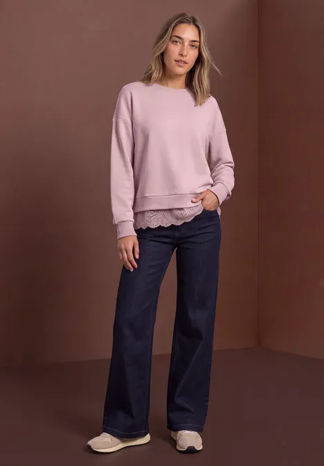 Sweat-shirt ample avec bordure en dentelle quiet rose Frau in rosa Sweatshirt und weiten Jeans steht vor braunem Hintergrund, lässige Pose mit Händen in Taschen.
