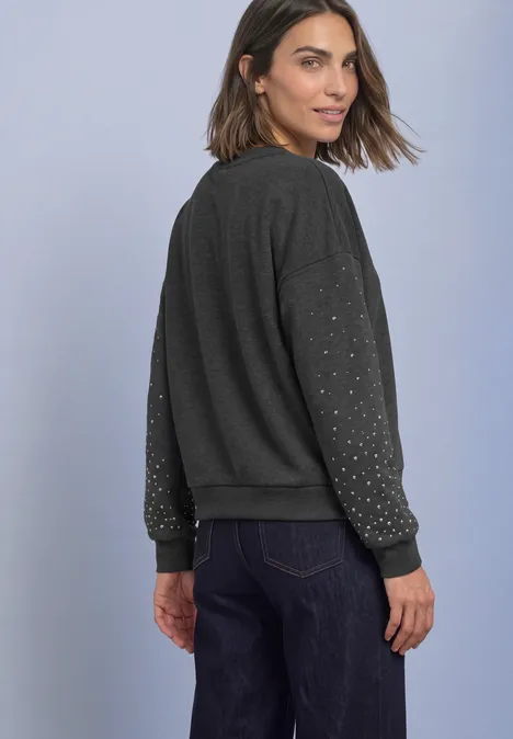 Sweat-shirt ample avec des rivets armor grey mel. Frau schaut über die Schulter, trägt dunkelgrauen Pullover mit Nietenärmeln und Jeans vor hellem Hintergrund.