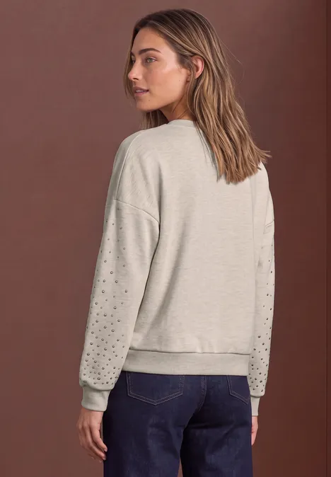 Rückansicht einer Frau in grauem Sweatshirt mit Lochmuster an den Ärmeln und blauen Jeans vor braunem Hintergrund.