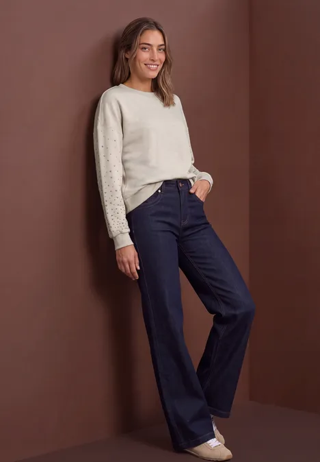 Frau in cremefarbenem Pullover und dunklen, weiten Jeans lehnt lässig an einer Wand.