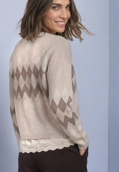 Dame in beiger Strickjacke mit Rautenmuster lächelt über die Schulter, darunter Spitzenbluse.