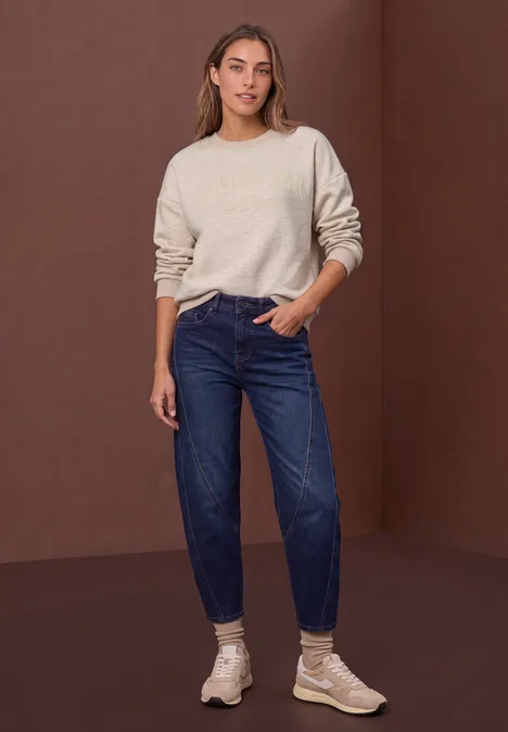 Loose Fit Sweatshirt mit Glitzer clay sand mel. Frau in beigem Pullover, locker sitzender blauer Jeans und hellen Sneakern vor schlichtem braunem Hintergrund stehend.