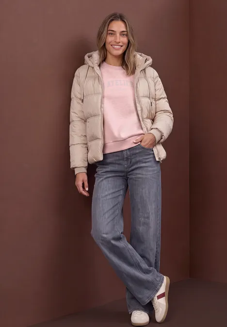 Loose Fit Sweatshirt mit Glitzer quiet rose mel. Frau in heller Steppjacke, rosa Pullover und blauen Jeans steht entspannt und lächelt.