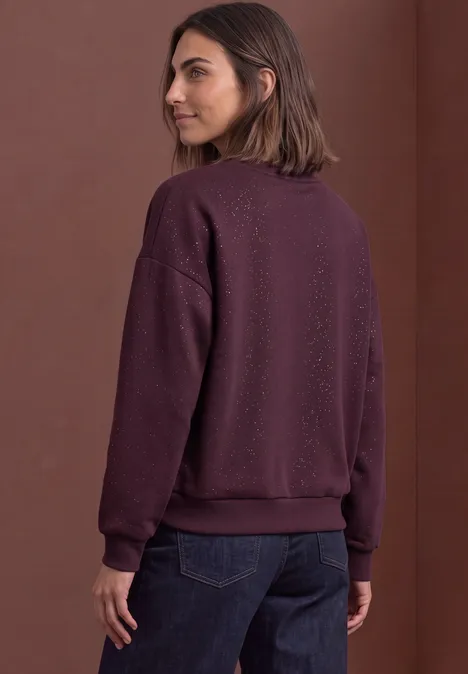 Loose Fit Sweatshirt mit Glitzer berry plum Frau mit mittellangen braunen Haaren in lila Sweatshirt und dunklen Jeans, seitlich von hinten aufgenommen.