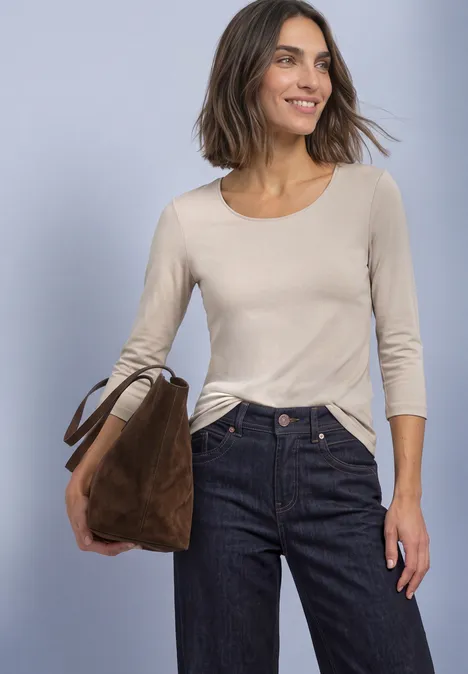 Une femme porte un top beige à manches longues, un jean foncé et tient un sac marron.