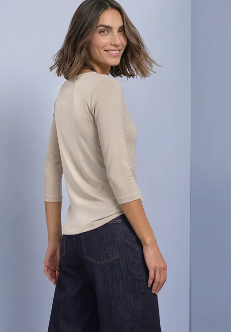 Femme vue de dos, vêtue d'un haut beige à manches 3/4 et d'un jean sombre, souriant en se retournant.