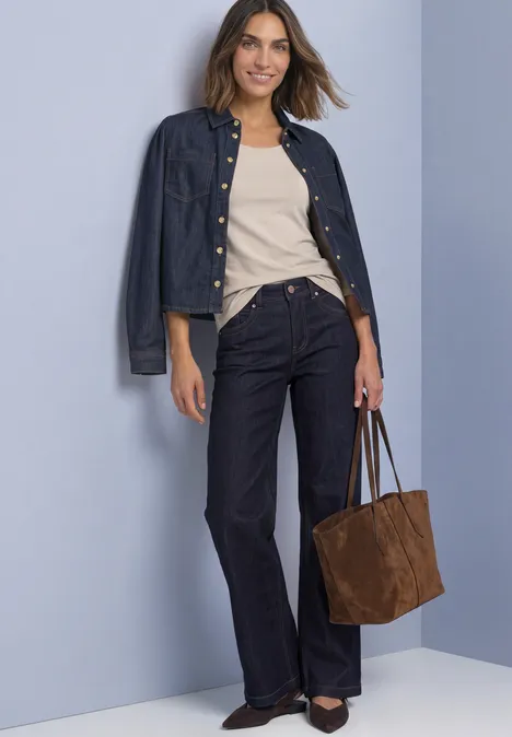 Femme en veste en jean, haut beige, jean flare foncé, chaussures noires et sac brun devant un mur clair.
