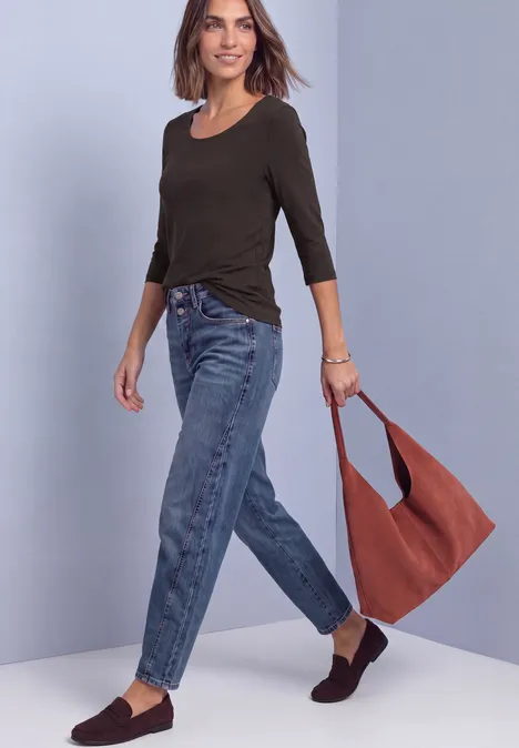 Frau in dunkelgrünem Langarmshirt, Jeans und braunen Slippern, die eine rostrote Schultertasche trägt.