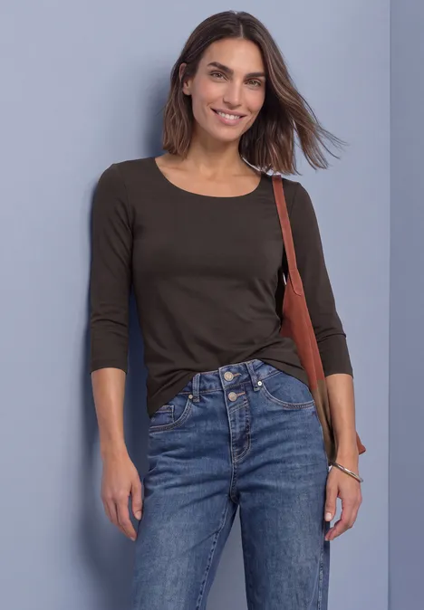 Eine Frau in schwarzem 3/4-Ärmel-Shirt und blauen Jeans mit Tasche, lächelnd vor grauem Hintergrund.