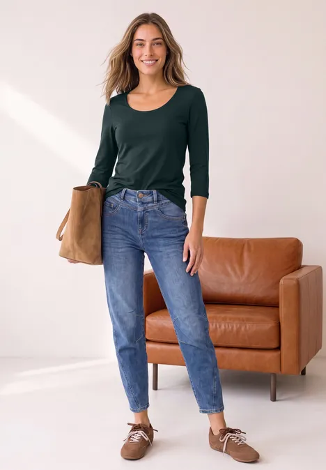 Jeune femme souriante dans un salon lumineux, portant un top vert, un jean bleu, des chaussures et tenant un sac.