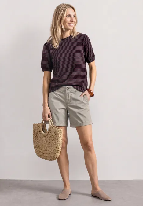 Frau in legerem Outfit: dunkelviolettes T-Shirt, graue Shorts, Basttasche und flache Schuhe.