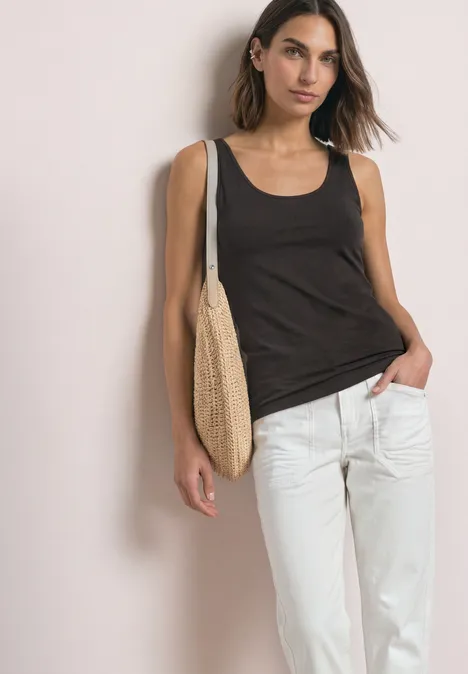 Jonge vrouw voor neutrale achtergrond, draagt een zwart topje, witte broek en een strotas.
