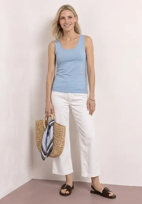Vrouw met lichtblauw hemdje, witte culotte, zwarte sandalen en rieten tas met sjaal, witte achtergrond.