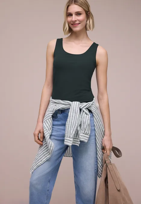 Basis top deep bound green Vrouw met zwart hemdje, blauwe jeans, gestreepte trui om de taille geknoopt en beige tas.