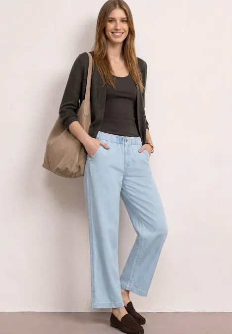 Vrouw in casual outfit: zwart topje, grijs vest, lichte wijde jeans, bruine loafers en een shopper tas.