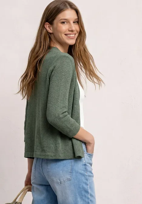 Femme vue de profil portant un gilet vert, un top blanc et un jean.