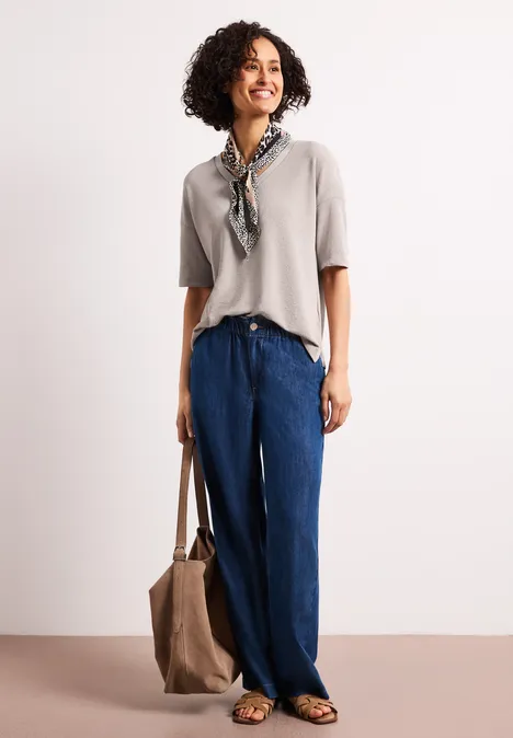 Vrouw voor witte achtergrond, draagt beige top, wijde blauwe broek, sjaal en tas