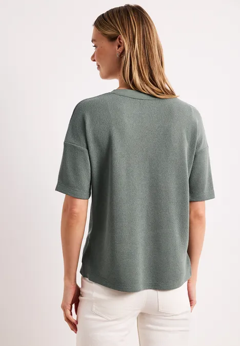 Vrouw van achteren met groen T-shirt met korte mouwen en witte broek tegen een witte achtergrond.