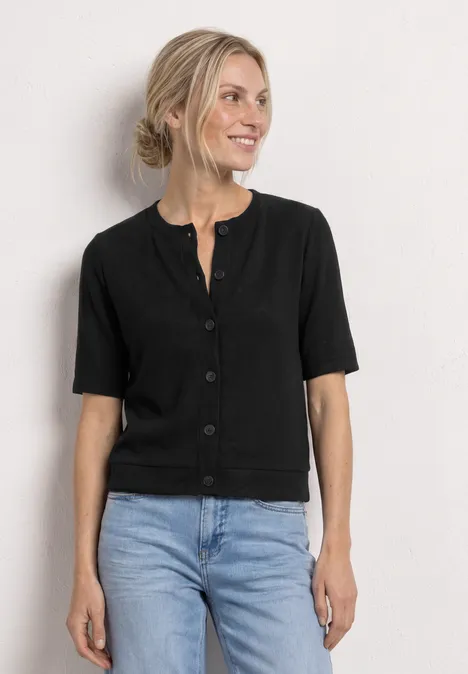 Eine Frau trägt eine schwarze Kurzarm-Strickjacke mit Knöpfen und blaue Jeans, vor neutralem Hintergrund.