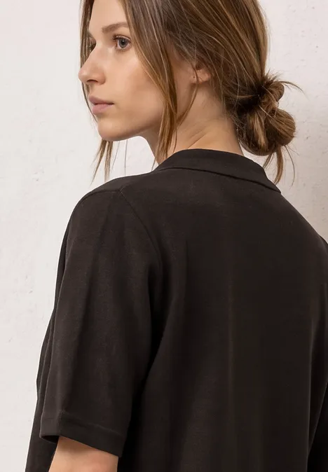 Junge Frau mit zusammengebundenem Haar trägt ein schwarzes Kurzarmshirt und blickt seitlich.