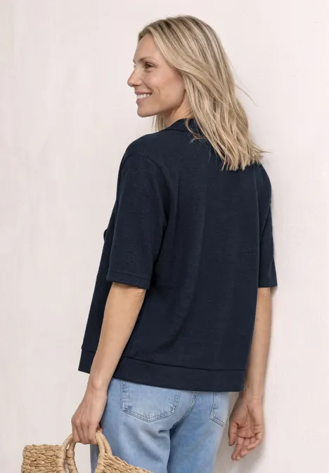 Person von hinten, trägt dunkelblauen Kurzarm-Pullover und helle Jeans, vor schlichtem Hintergrund.