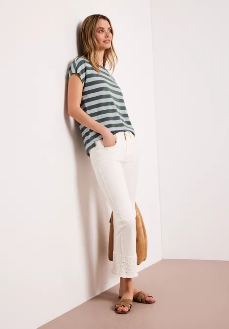 Shirt met hangende schouders, strepen en glitter bound green Vrouw leunt tegen muur, draagt groen-wit gestreept shirt, witte flared broek met franjes en sandalen.