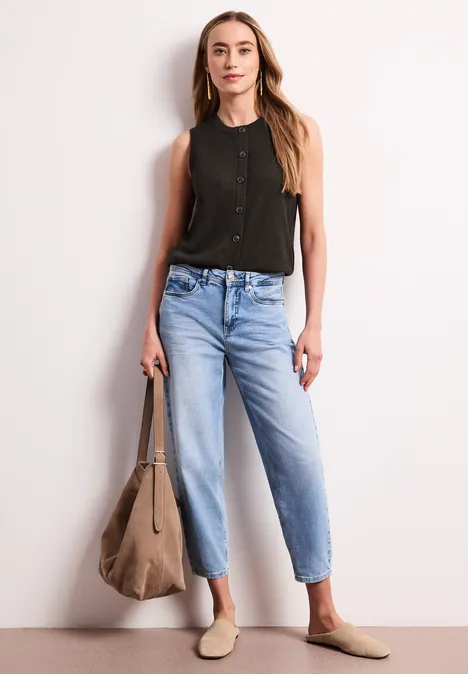 Frau trägt schwarzes ärmelloses Top, helle Jeans, beigetönige Slipper und große Tasche vor weißem Hintergrund.