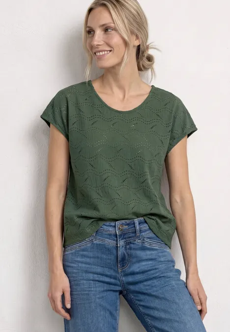 Vrouw in een groen t-shirt en blauwe jeans tegen een neutrale achtergrond.